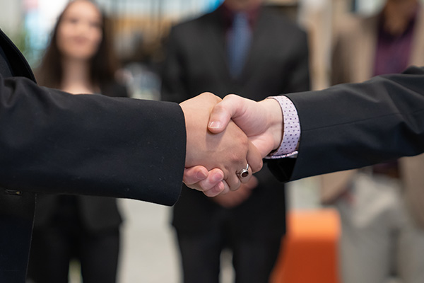 close up of a handshake with business students standing in the background