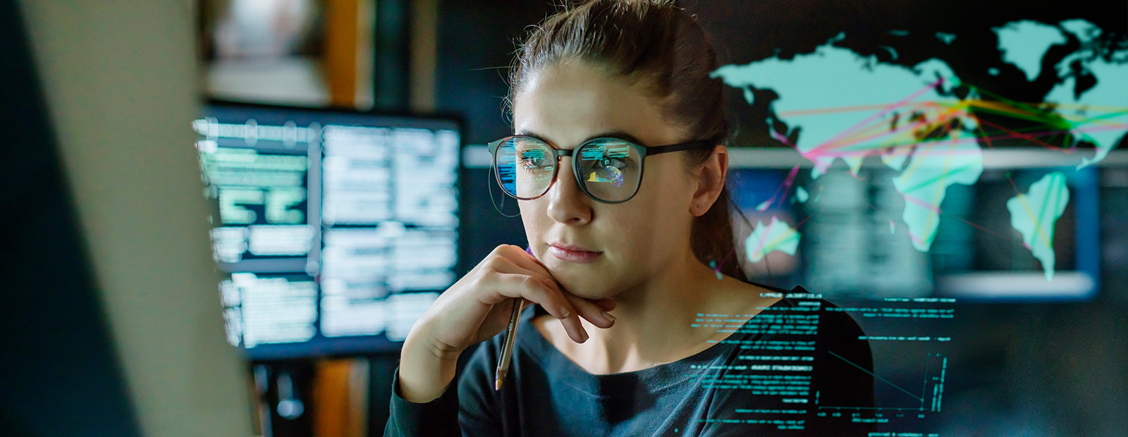 young woman wearing glasses surrounded by computer monitors in a dark office