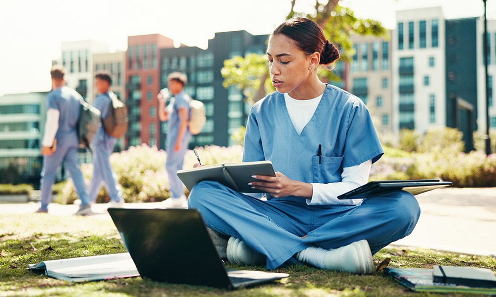 nursing student sitting outside of campus buildings with nursing students walking behind her