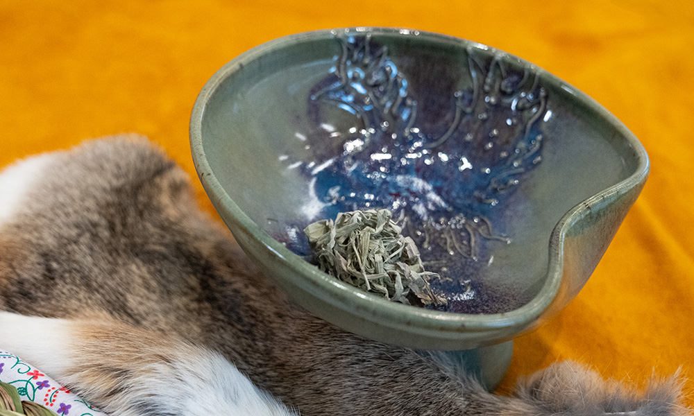 A smudge bowl filled with sage, resting on a mat.