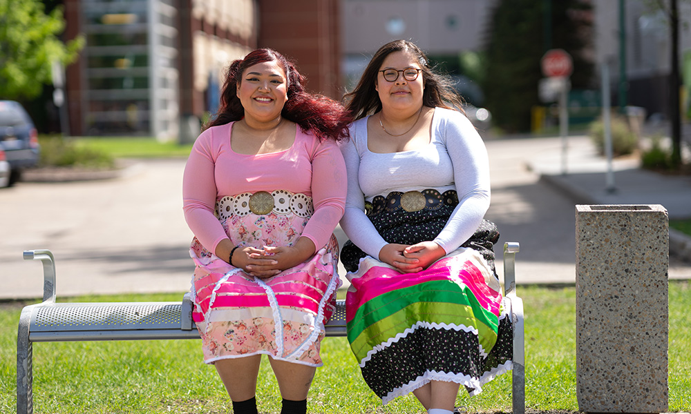 Two Indigenous students wearing ribbon skirts sit side by side on a bench outdoors, smiling in the sunlight.