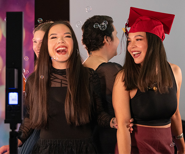 Two graduating students laugh and celebrate together, one wearing a red cap and gown, surrounded by friends.