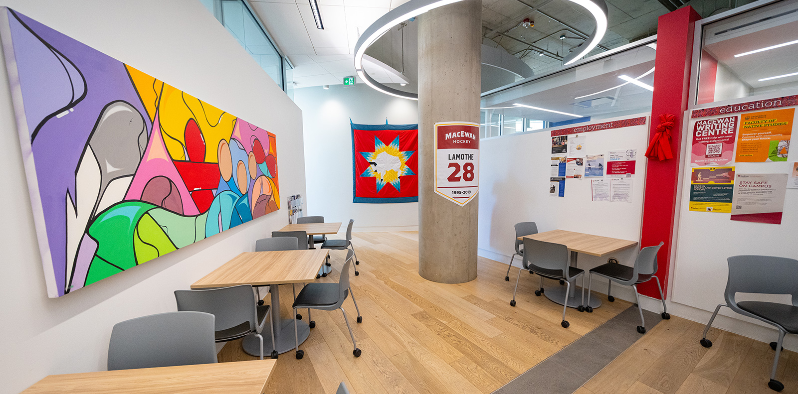 Tables and chairs line a bright hallway at kihêw waciston, with colourful student artwork on the walls