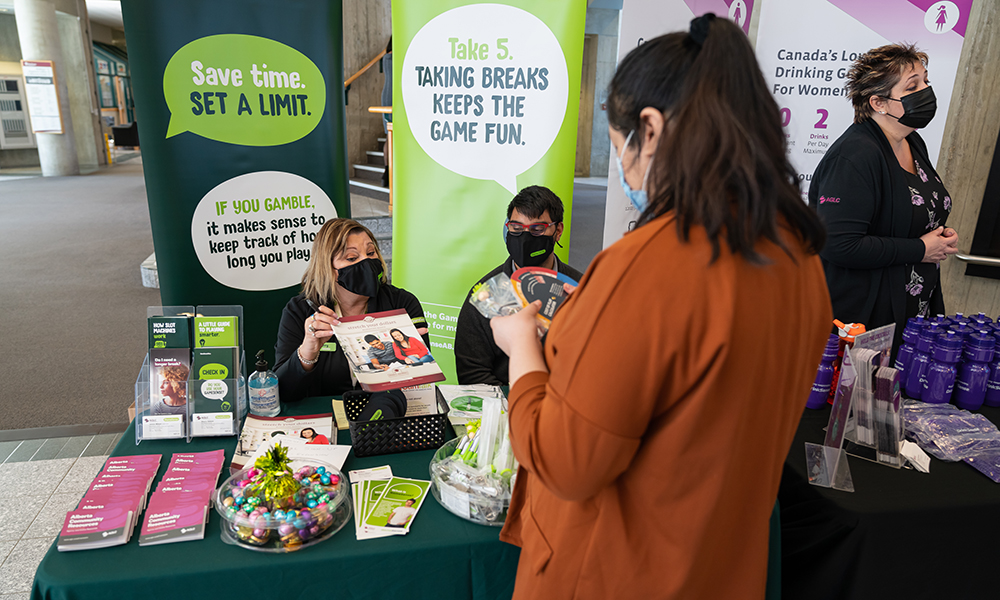 Two students seated at an information booth provide paper materials and talk to another student, who is standing in front of the booth.