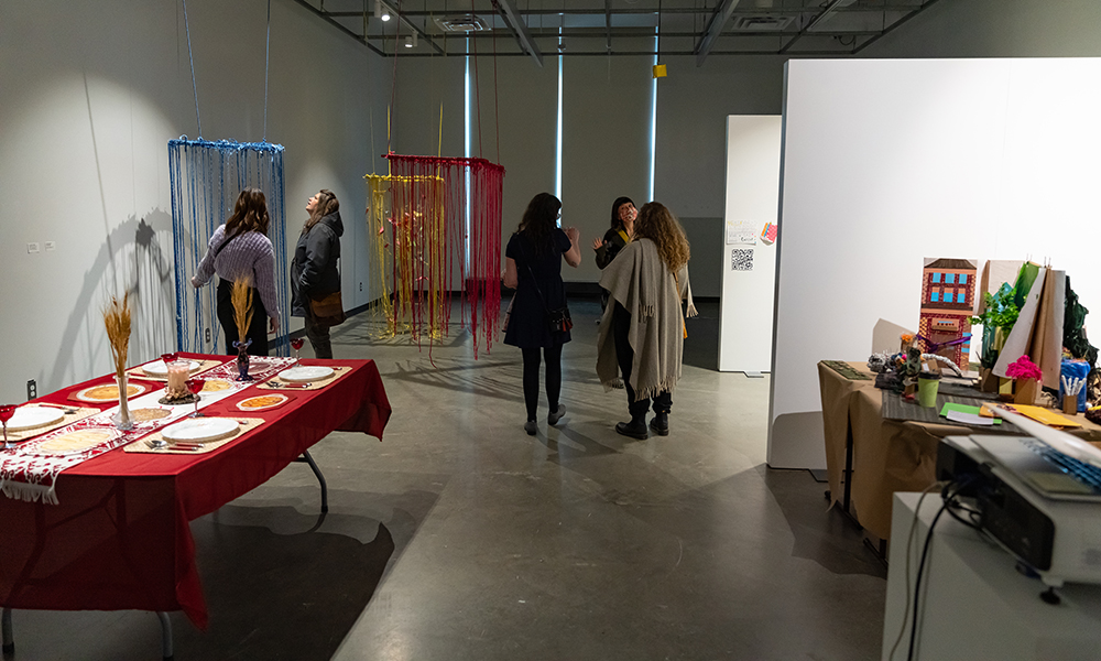 People gather around art installations in an art gallery, including an installation of fabric strings suspended from the ceiling and a table full of plates and vases.