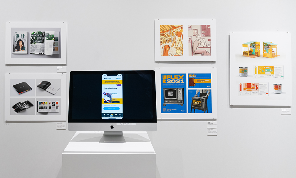 Samples of graphic design work hang on the white walls of an art gallery, and a large computer monitor with graphics on the screen sits on a stand in the middle.