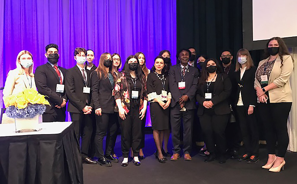 A group of students in business attire stand together on a stage in front of a purple curtain.