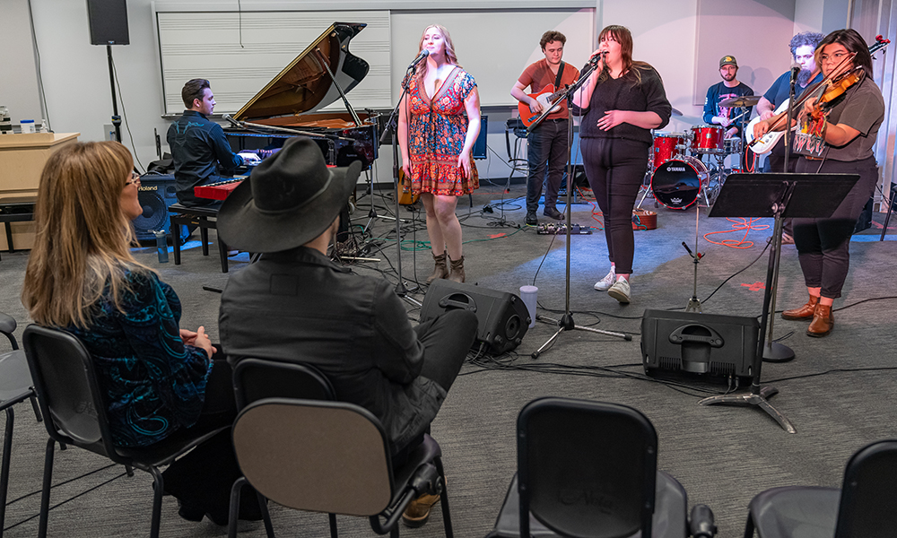 Students sing into microphones in a classroom while country singers Brett Kissel and Michelle Wright watch them while seated in a row of chairs.