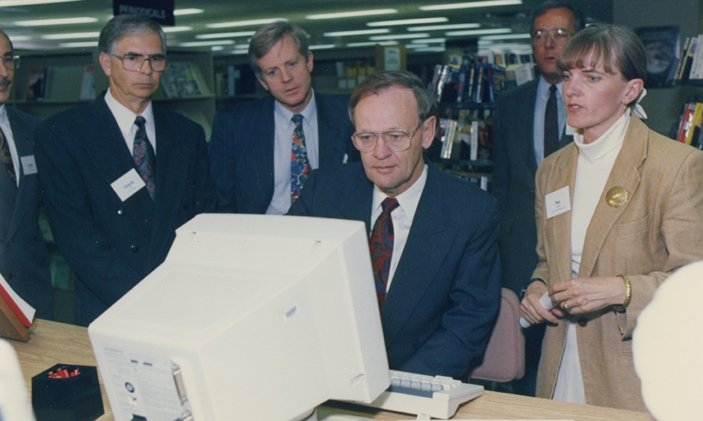 A group of people, including former prime minister Jean Chretien, gather in front of an old computer monitor.