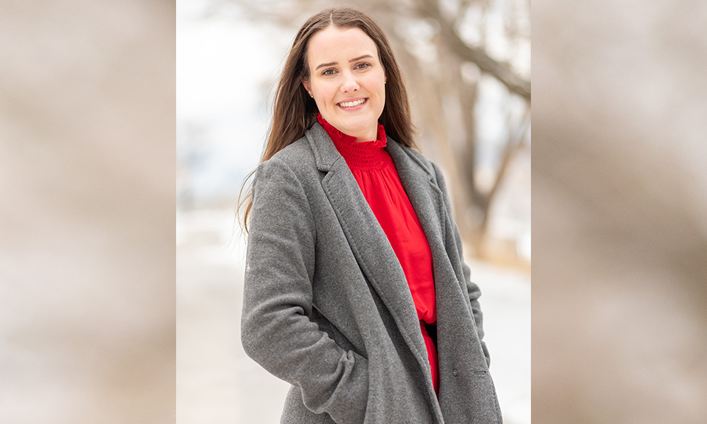 Becca Willson stands outside on a chilly looking day, wearing a grey coat. There are bare, leafless trees behind her.