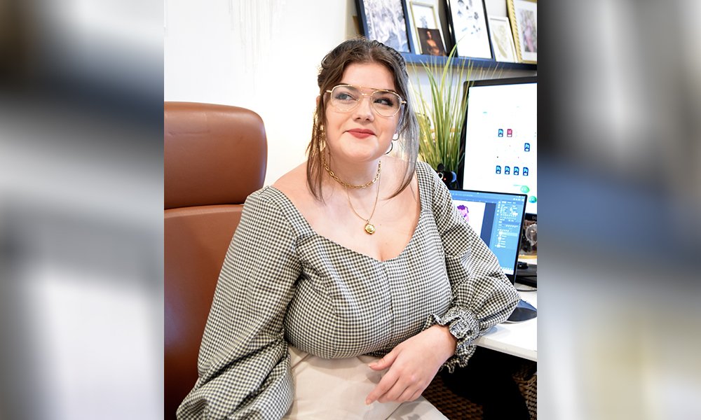 Shae McMullin sits on a brown leather chair in front of a desk with a computer on it.