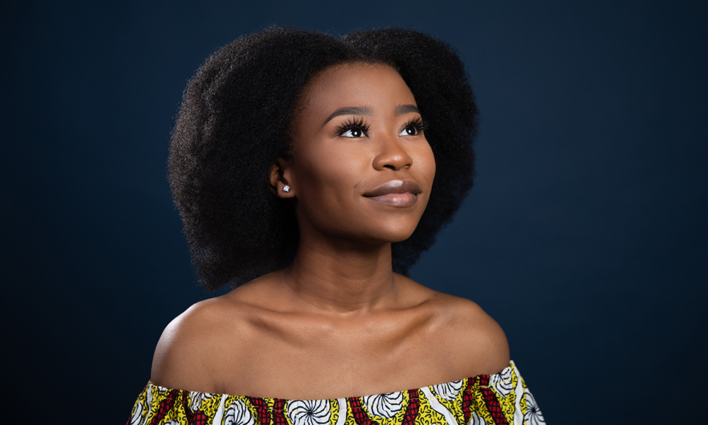 Sokhana Mfenyana is wearing a bold patterned shirt and gazing off into the distance, as she sits against a dark background.