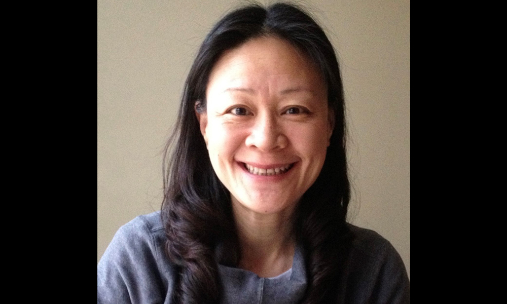 Xiaomei Yang is standing against a beige background. She is smiling and wearing a blue top.