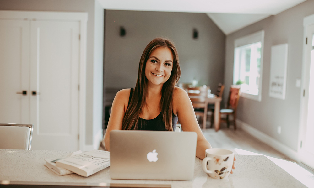 Karina Laitres sits at a desk inside her home, with a laptop and a coffee mug on the desk i front of her.