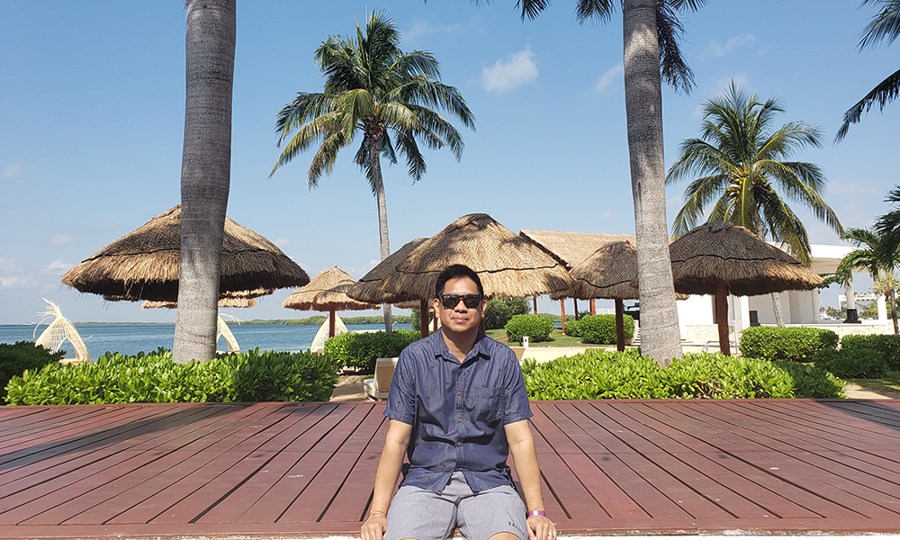 Larry Garcia sits outside on a sunny day, with palm trees behind him.