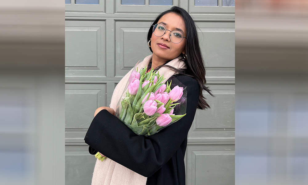 Agnya Patel stands outside wearing a long beige scarf, holding a bouquet of pink flowers.