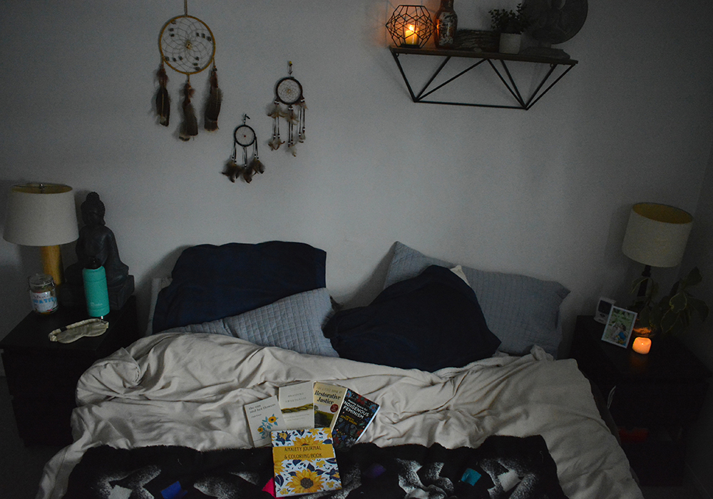 Photo of a messy bed with books on it. Three dreamcatchers hang on the wall behind the bed.