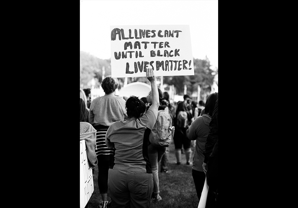 Black and white photo of protesters. One is holding a sign that says "All lives can't matter until Black lives matter!"