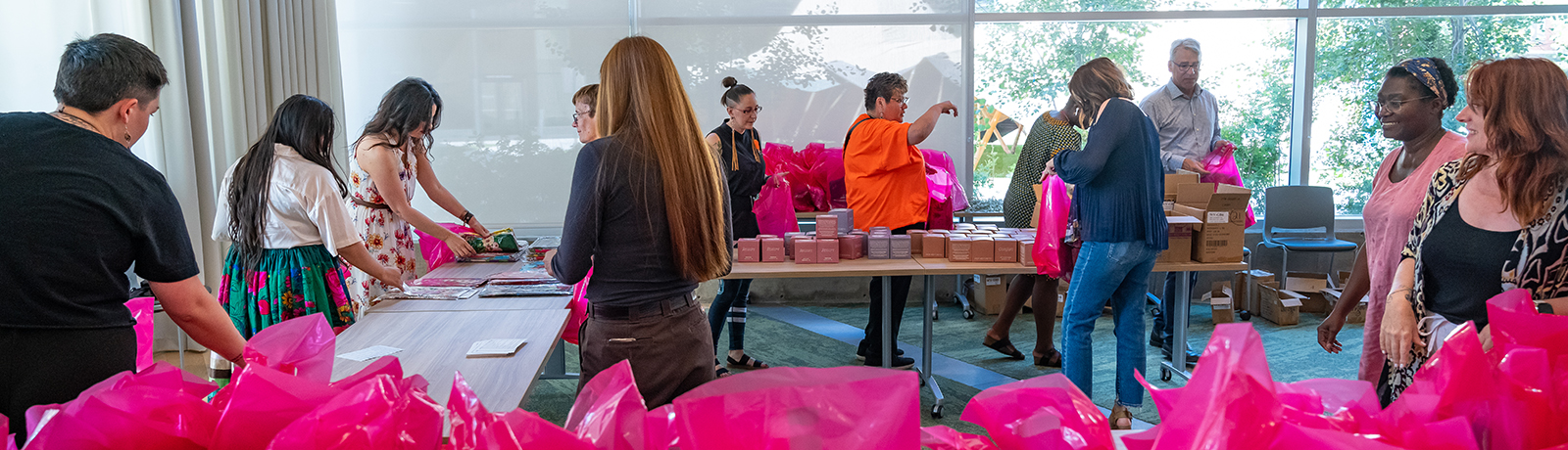 A group of volunteers gather around a large table, placing objects in pink plastic bags.