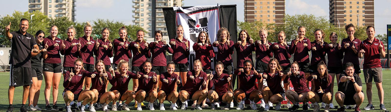 The players and coaches from the 2021 women's Grffins soccer team stand in a row. They are each holding out one hand to display their championships rings, and making a number one gesture with the other hand.