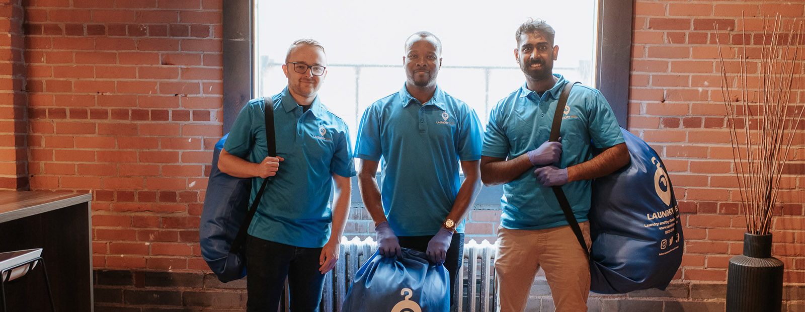 Andrew Bartholet, Akinwale Ajayi and Dhan Moonian stand in front of a brick wall wearing LaundryWell shirts and holding laundry bags