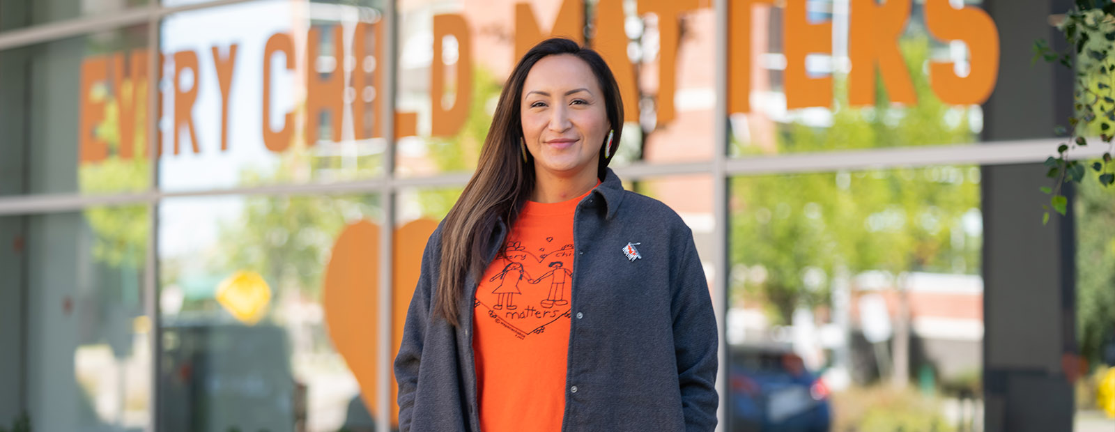 Terri Cardinal wears an orange shirt and stands in front of the glass windows of kihêw waciston, the university's Indigenous centre