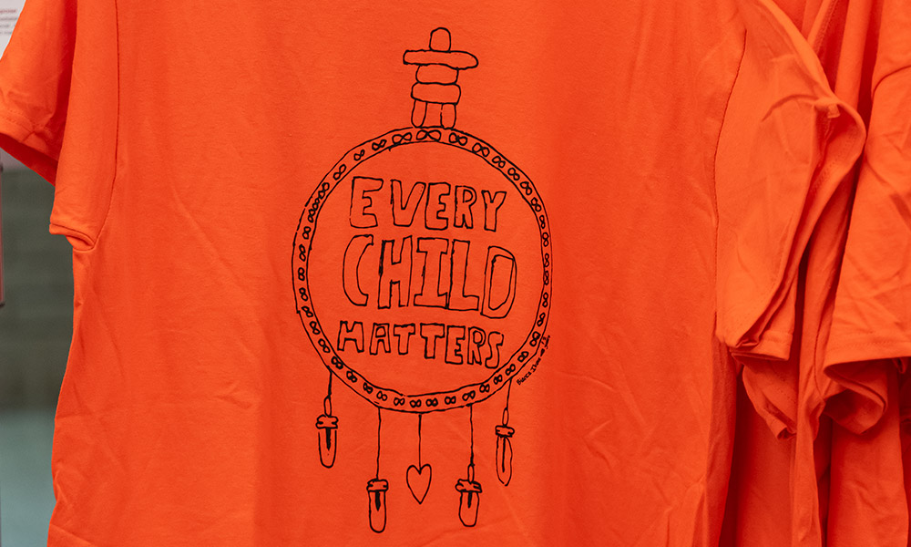 An Orange Shirt Day t-shirt design with a stylized dreamcatcher that includes an inukshuk, the infinity symbol from the Metis flag and a small heart.