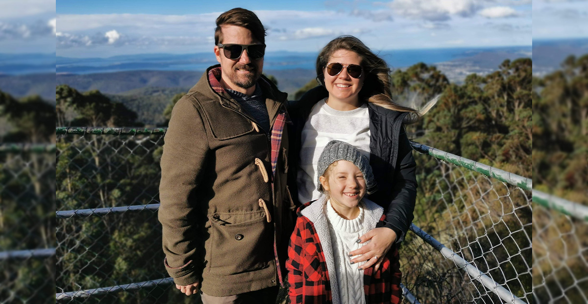 Angela Merriott and her fiancé and daughter stand before a tree canopy