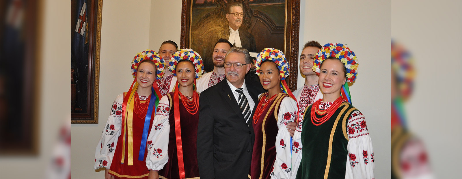 Gene Zwozdesksy stands in front of his portrait flanked by Shumka dancers dressed in brightly coloured dresses and head pieces