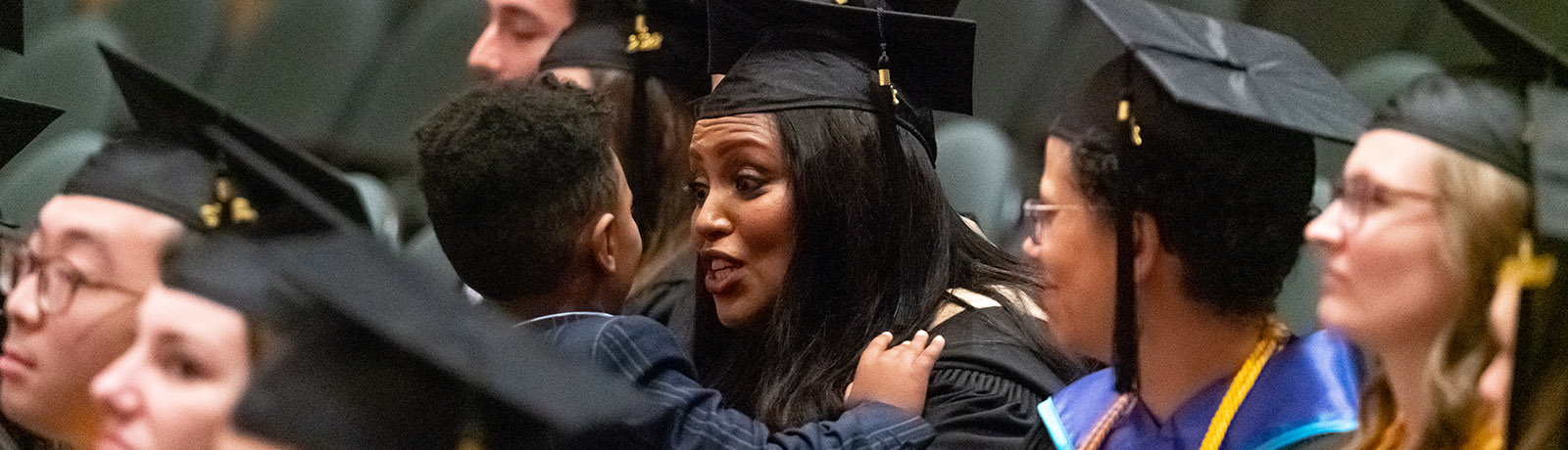 A graduate holds a small child and speaks to him while seating with other graduates during fall convocation