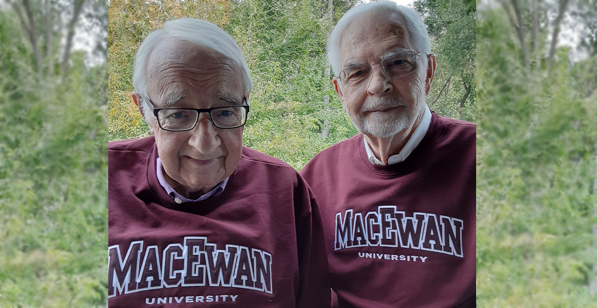 Dr. Terry Flannigan and Dr. Chuck Day stand together wearing MacEwan sweatshirts in front of a background of green trees.