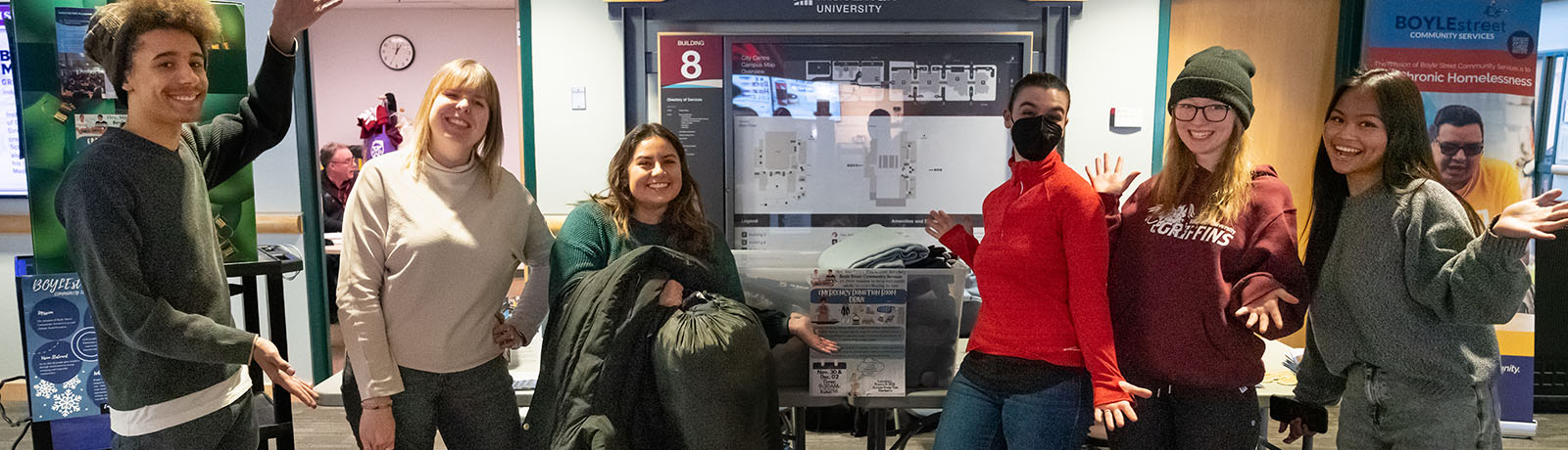 Students in INTD 205 stand in from of a table in Building 8 with bins to collect warm winter clothing.