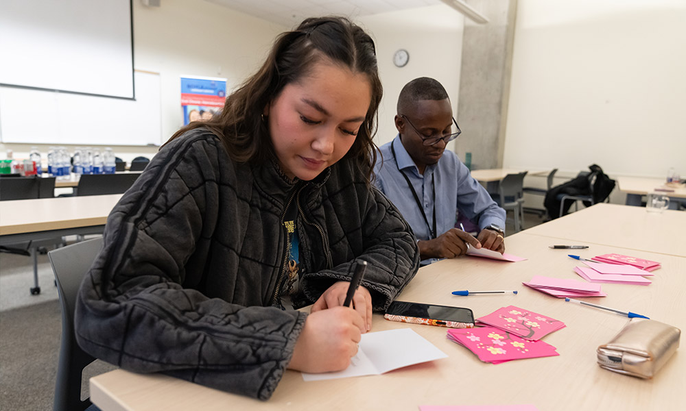 Volunteers sit at a table in a classroom writing cards to include in the kits