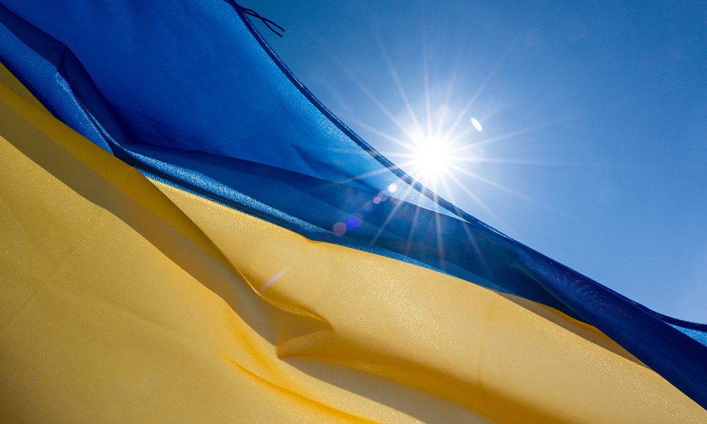 A Ukrainian flag flies in the wind with a bright sun and blue sky in the background