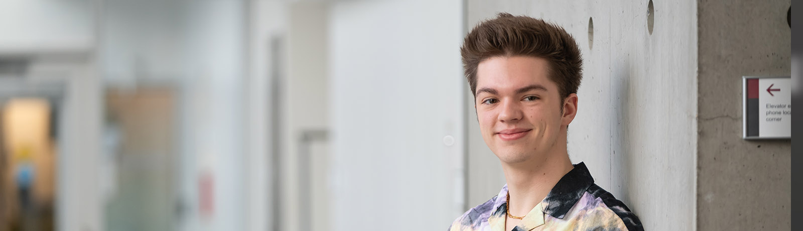 Brayden Ritter wears a black and yellow patterned shirt and leans against a wall in a hallway on campus