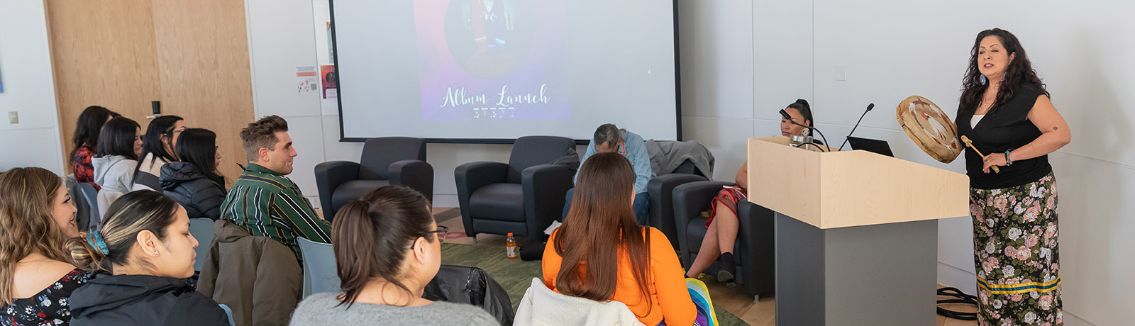 A group of people sit in an audience while a women holds a drum and speaks at the launch event