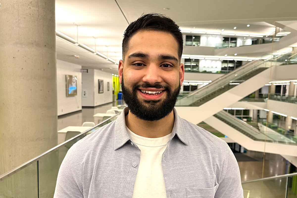 Harnoor Dhaliwal is wearing a white shirt and grey overshirt, and smiles at the camera while standing in Allard Hall