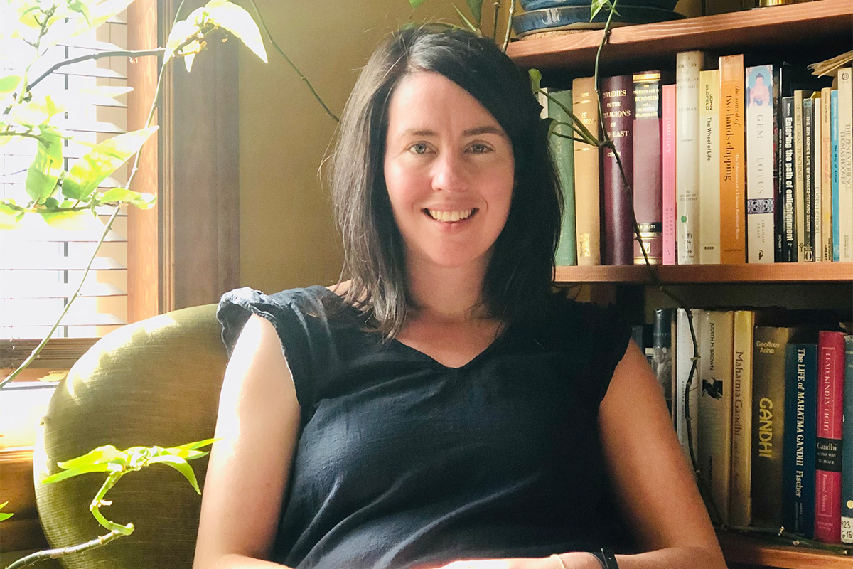 Lindsey Whitson is wearing a black shirt and is sitting on a green chair next to a bookshelf. She smiles at the camera