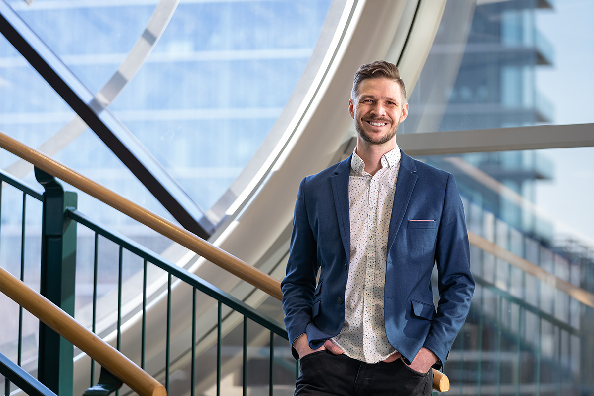 Shaun Bosch wears a white shirt with black dots beneath a blue blazer. He stands in front of a window in the library and smiles at the camera