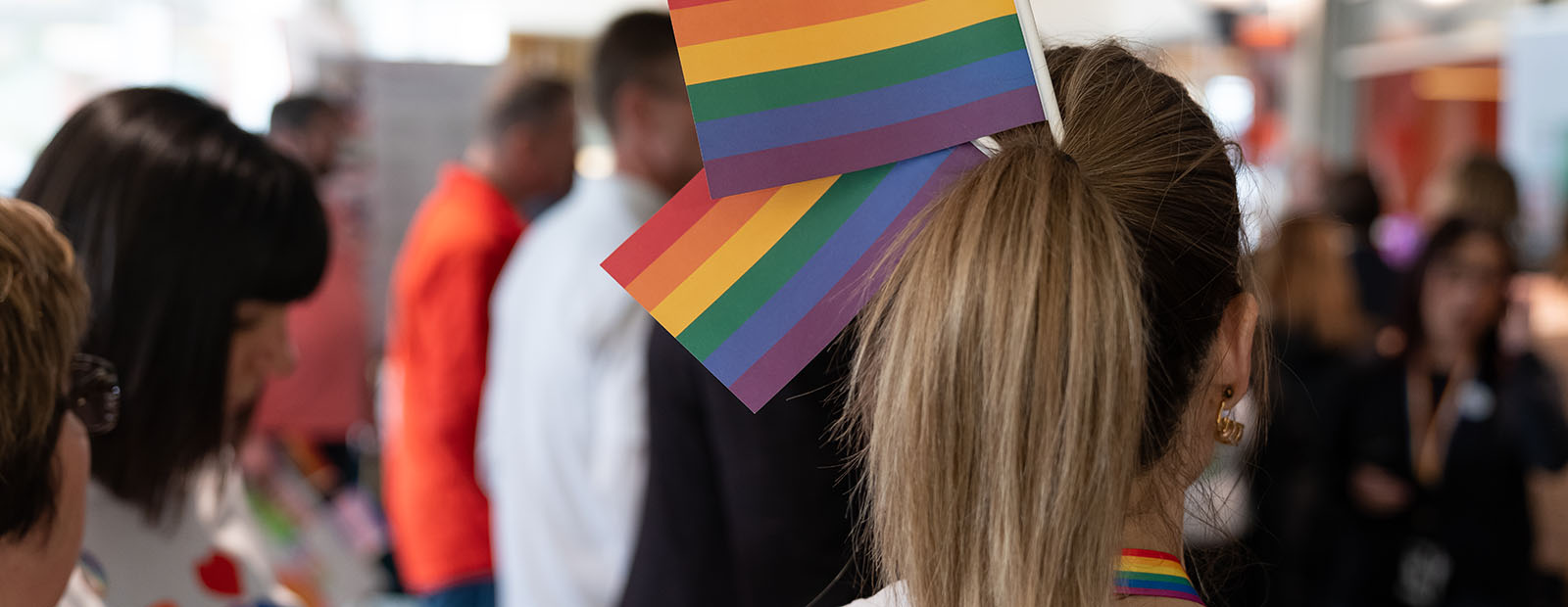 A group of people gather for a Pride Week event. At the centre of the photo is a woman with Pride flags tucked into her ponytail.