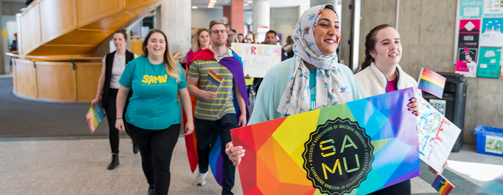 Students walk through Building 6 carrying rainbow flags in a campus Pride march, all smiling