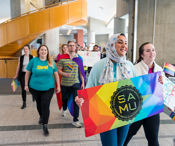 A group of students walk through Building 6 as part of a Pride March, smiling and waving rainbow flags