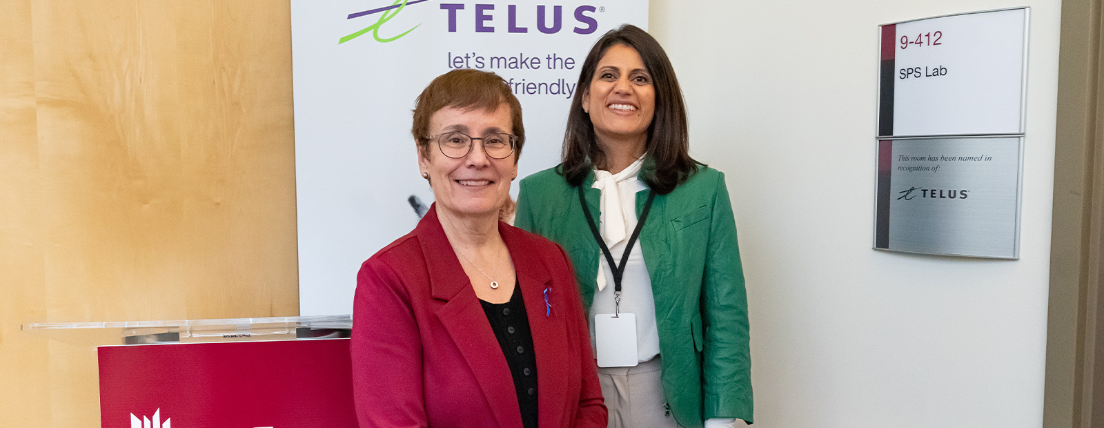 Dr. Annette Trimbee and Zainul Mawji stand outside of a closed classroom door. A plaque beside the door notes that the room is named for TELUS