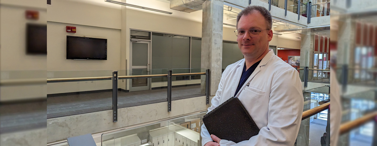 Jason Kossowan stands in a hallway in Building 9, wearing a lab coat and holding a clipboard.
