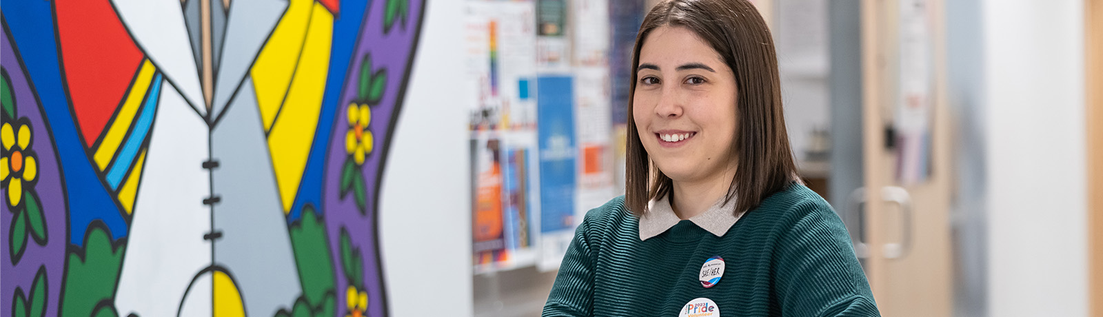 Michelle Viveiros stands beside a colourful mural in the Centre for Sexual and Gender Diversity wearing a green sweater and a Pride button