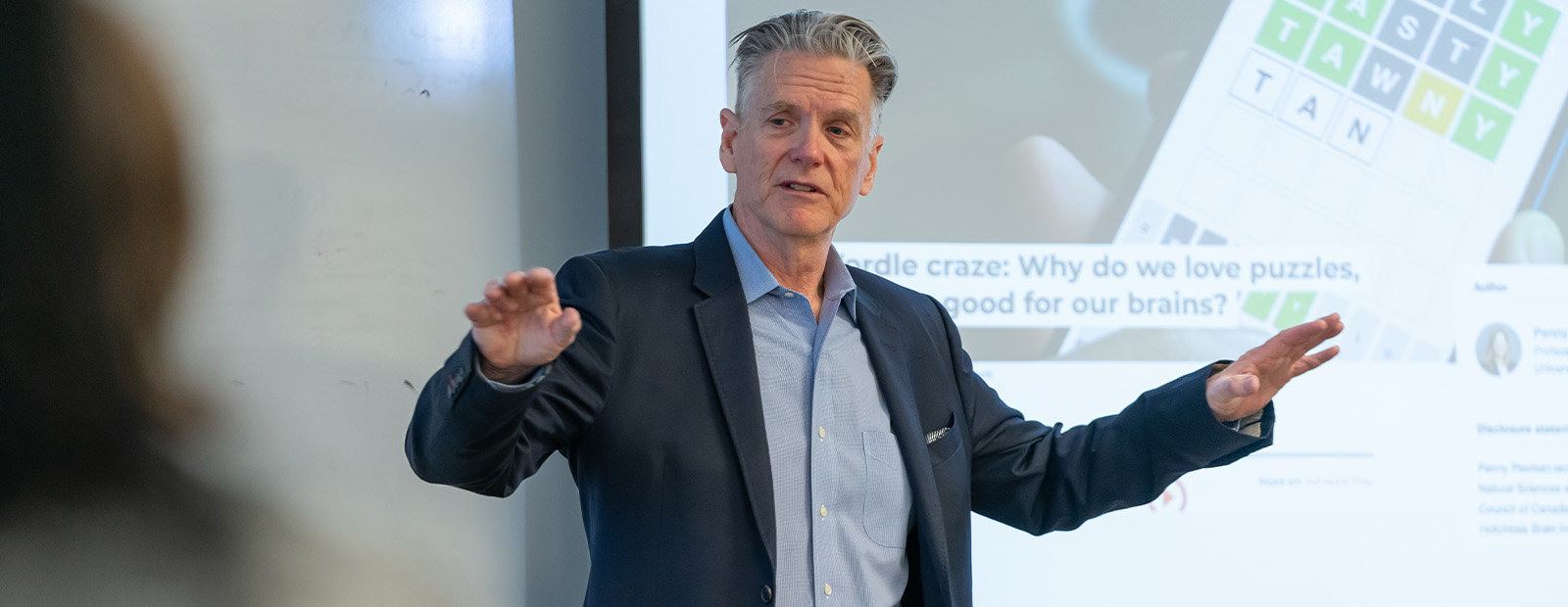 Scott White stands at a podium in a MacEwan classroom and gestures outwardly. A slideshow behind him shows a World puzzle