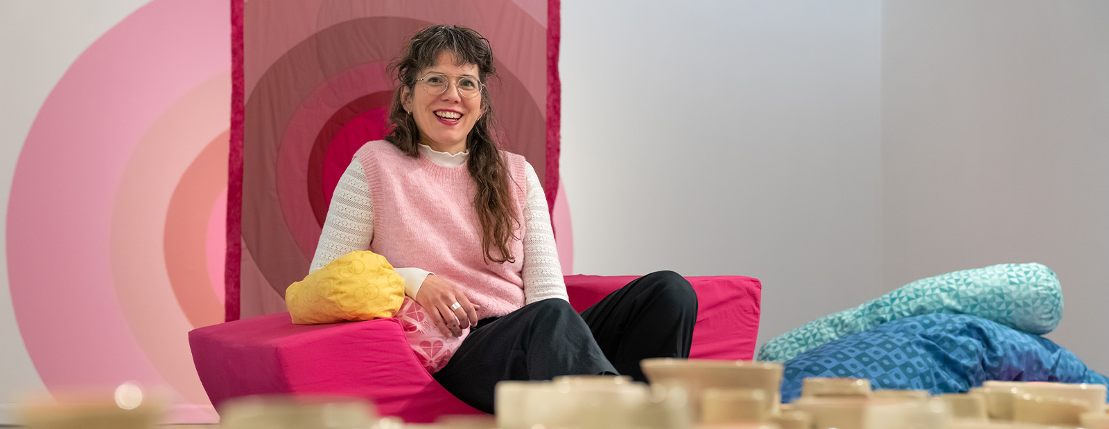 Kerri-Lynn Reeves sits on a bright pink semi-circular cushion in front of a backdrop of pink and white concentric circles. Dozens of ceramic vessels sit on a table in front of her