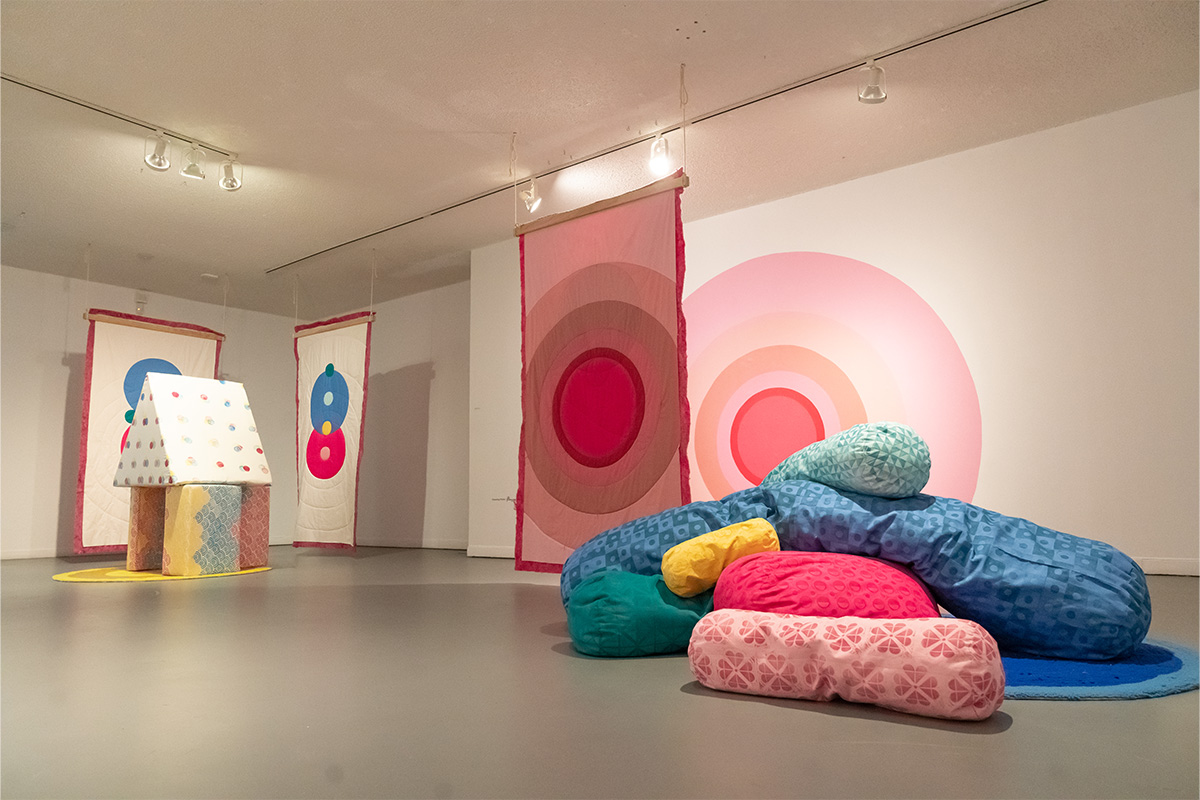 A number of soft cushions in varying shades of pink and white lay on the floor, and others are arranged to create a structure of a house. The wall behind is painted with pink and white concentric circles