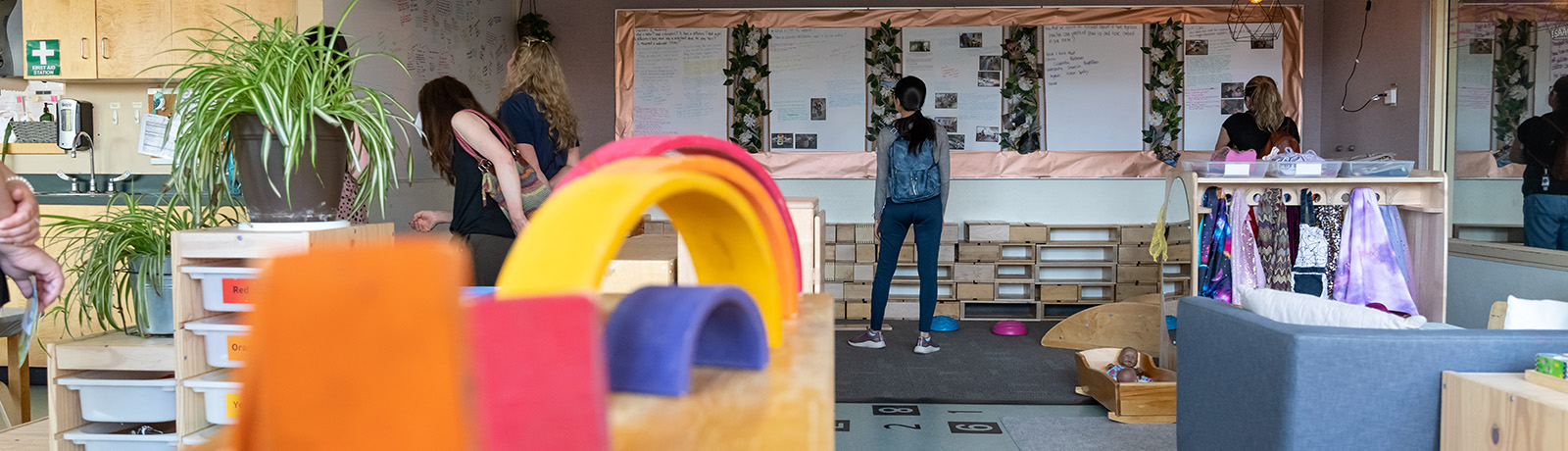 A wide angle photo of one of the rooms in ELM with colourful fainbow toys in the foreground, people milling around and children's artwork hanging on a string from the ceiling.
