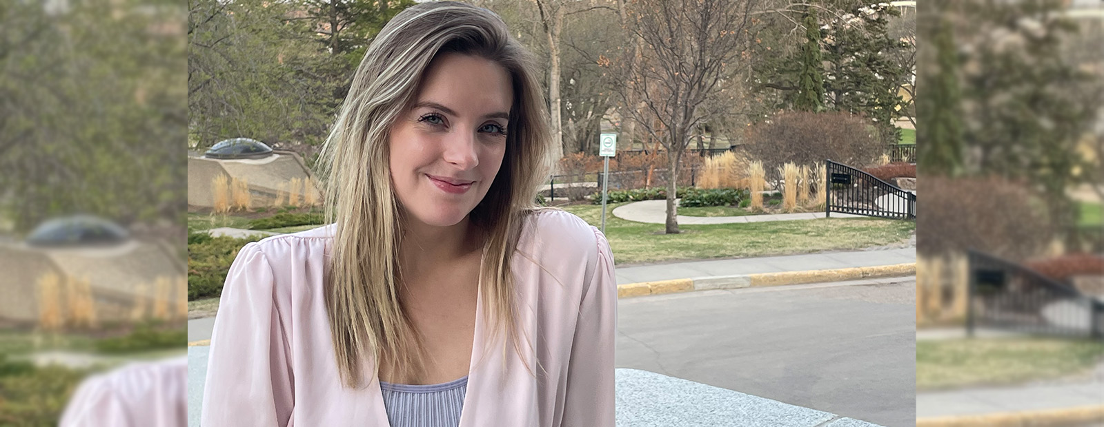 Olivia Hardy stands in front of green park space, wearing a pink cardigan and smiling at the camera.